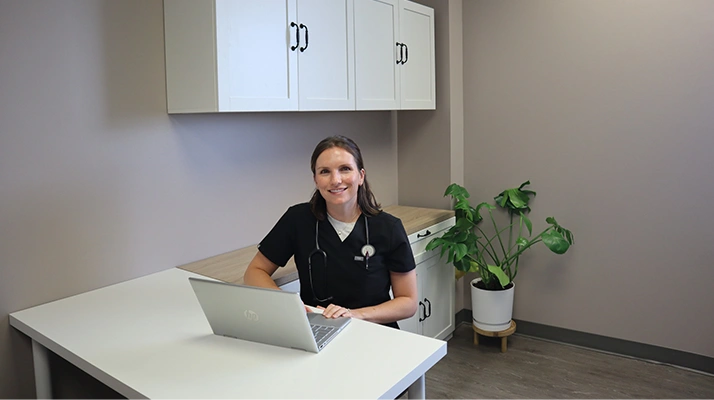 Chiropractor Towson MD Caroline Chell sits at a desk using a laptop in a clean, modern clinic setting, with a potted plant and cabinets visible in the background.