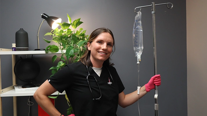 Caroline Chell stands beside an IV drip setup in a clinical room, holding the IV line while wearing medical gloves.