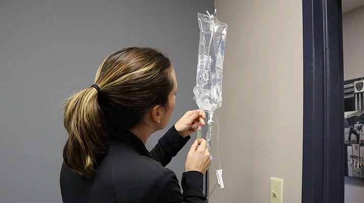 Caroline Chell adjusts an IV infusion bag mounted on a stand, preparing or monitoring a treatment in a clinical setting.